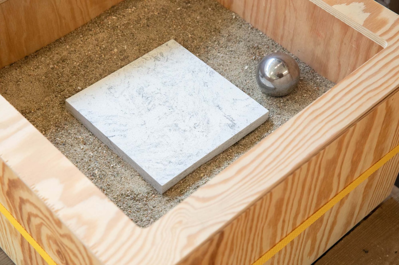 A square piece of LOCKEBO worktop in a wooden box filled with sand beside a metal ball used.
