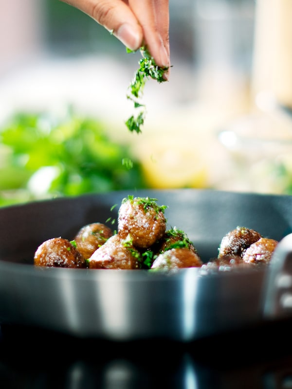 A person sprinkles chopped green herbs on to HUVUDROLL plant balls in a IKEA 365+ frying pan on a hob.