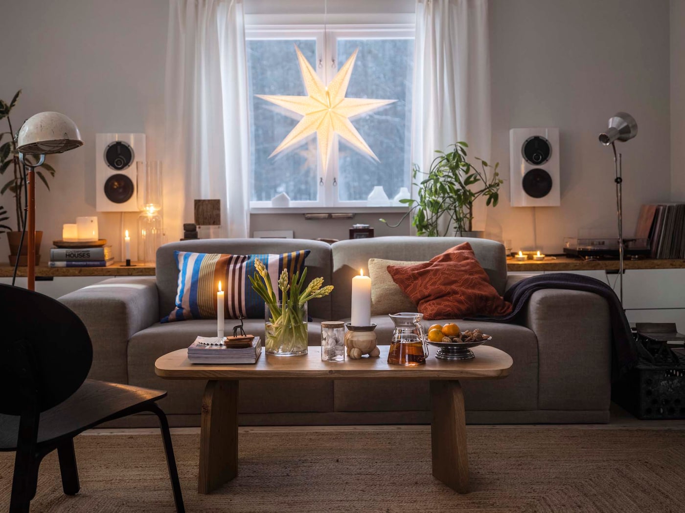 Cosy, dimly lit living room with a wide sofa, coffee table decked with candles and flowers, and a paper star hanging in the window in the background.