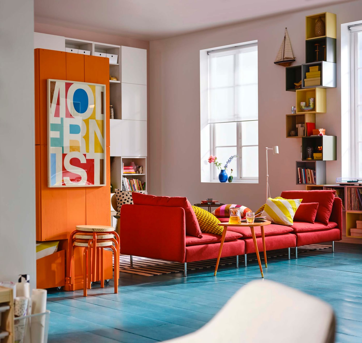 A room with a blue floor. Orange cupboards and stools stand alongside a red modular sofa.