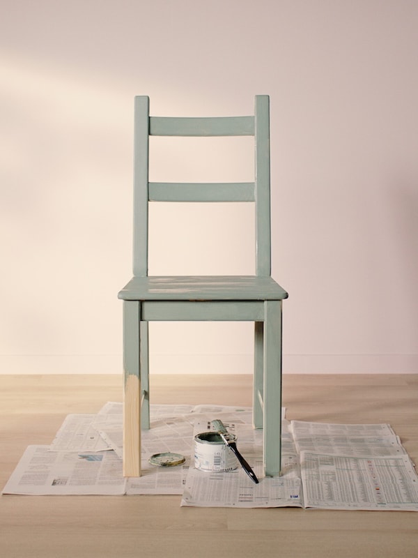 Židle IVAR natíraná světle zelenou barvou stojící na starých novinách v prázdné místnosti se světlou dřevěnou podlahou a světle růžovými stěnami.